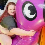 A person with red hair and a dark top sits smiling on a large purple inflatable toy shaped like an animal, indoors with a yellow curtain and decorative lights in the background.