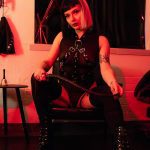 A woman with a black and blonde bob sits confidently in a chair, wearing black harness attire and holding a riding crop, in a dimly lit room with red lighting.