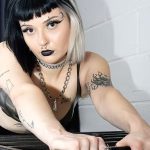 A person with black and white hair, bold make-up, and tattoos leans forward on a metal structure, wearing a black outfit and chain necklace, in a room with white brick walls and dark curtains.